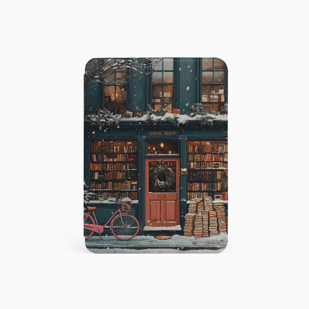 The Kindle case features a snowy bookstore with a red door and a black sign reading "Cherry." The exterior and roof are covered in books, highlighting the book-filled charm of the place. Warm lights glow around the store, and snowflakes gently fall from the sky, creating a peaceful and cozy atmosphere.