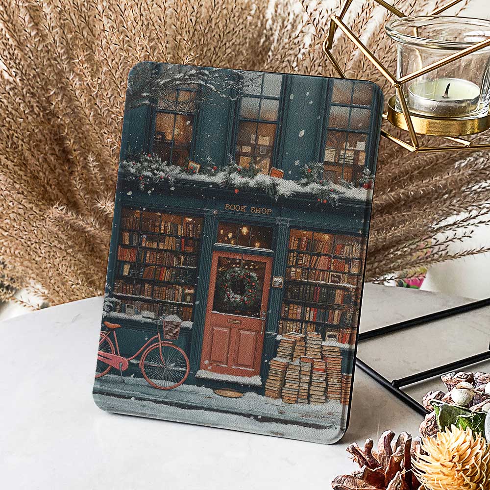 The Kindle case features a snowy bookstore with a red door and a black sign reading "Cherry." The exterior and roof are covered in books, highlighting the book-filled charm of the place. Warm lights glow around the store, and snowflakes gently fall from the sky, creating a peaceful and cozy atmosphere.