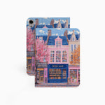 The iPad Pro M4 Case design features an illustration of a city street with vibrant buildings. The most prominent is a pink building in the center, labeled "BOOK SHOP." Large windows on the building display colorful bookshelves, creating an atmosphere full of knowledge and culture.