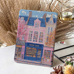 Design for a Kindle softcolor case featuring an illustration of a pink building with a blue roof and white-framed windows. In front, a bookshelf labeled 'BOOK SHOP' is displayed.
