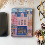Design for a Kindle softcolor case featuring an illustration of a pink building with a blue roof and white-framed windows. In front, a bookshelf labeled 'BOOK SHOP' is displayed.