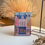 The iPad Pro M4 Case design features an illustration of a city street with vibrant buildings. The most prominent is a pink building in the center, labeled "BOOK SHOP." Large windows on the building display colorful bookshelves, creating an atmosphere full of knowledge and culture.