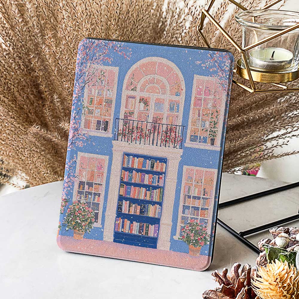 A kindle_colorsoft_case design featuring a bookstore with a blue door and an arched window showcasing bookshelves inside. The store has white windows on either side, also displaying books. Two green potted plants sit at the entrance, adding vibrancy. The overall color scheme of blue and pink creates a warm and peaceful atmosphere.