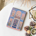 A kindle_colorsoft_case design featuring a bookstore with a blue door and an arched window showcasing bookshelves inside. The store has white windows on either side, also displaying books. Two green potted plants sit at the entrance, adding vibrancy. The overall color scheme of blue and pink creates a warm and peaceful atmosphere.