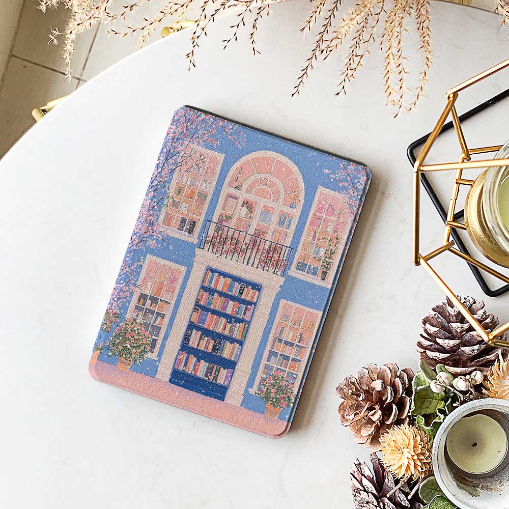 A kindle_colorsoft_case design featuring a bookstore with a blue door and an arched window showcasing bookshelves inside. The store has white windows on either side, also displaying books. Two green potted plants sit at the entrance, adding vibrancy. The overall color scheme of blue and pink creates a warm and peaceful atmosphere.