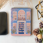 A kindle_colorsoft_case design featuring a bookstore with a blue door and an arched window showcasing bookshelves inside. The store has white windows on either side, also displaying books. Two green potted plants sit at the entrance, adding vibrancy. The overall color scheme of blue and pink creates a warm and peaceful atmosphere.